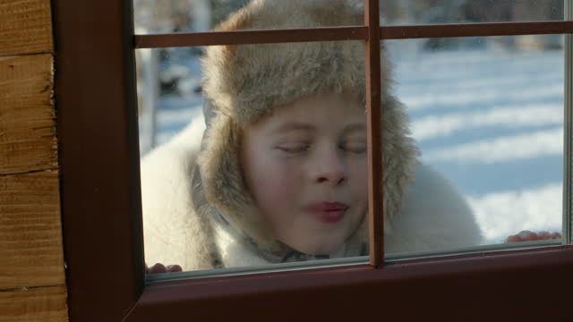 Child playing outdoors in winter snow peering into a wooden house through the glass window pane in a warm fir hat and jacket with a close up on the face