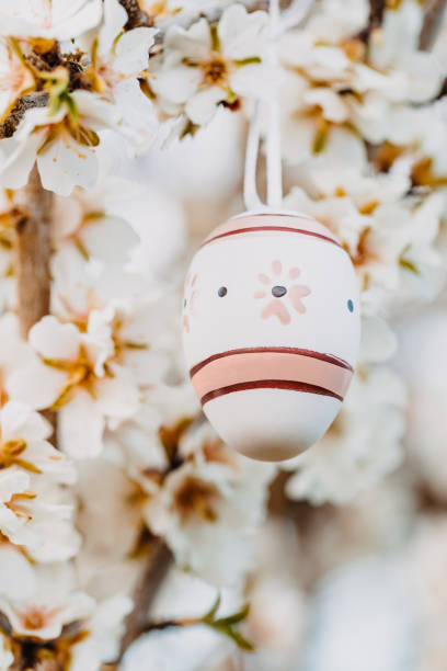 Close up of a beige boho easter egg hanging in between fragrant almond blossoms on an almond tree. Color editing with added grain. Very selective focus. Part of a series.
