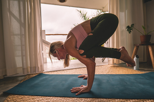 Beautiful woman exercising yoga at sunrise enjoying sea view from her balcony. People practicing sport indoors on yoga mat.