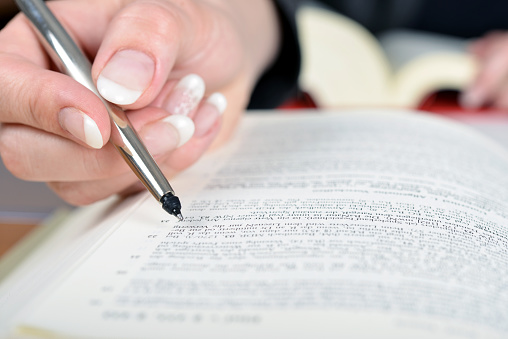 Close-up of a woman with pretty fingernails reading and researching in a book, marking the lines with a silver pen