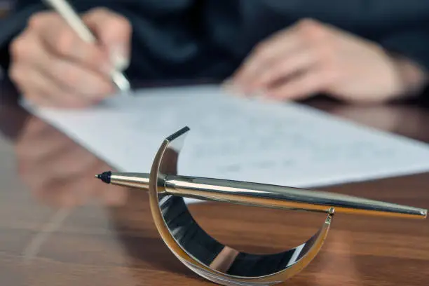 Female lawyer, out of focus in the background, in black robe works in her office with a penholder in the foreground