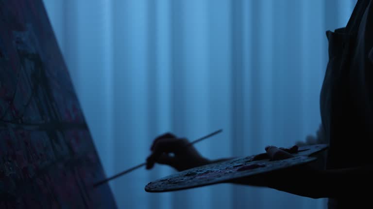 Silhouette of woman's hand draws a picture with oil paints on canvas with a brush. Female artist creates in an art workshop, applying strokes to a canvas. Modern art. Slow motion. Close up