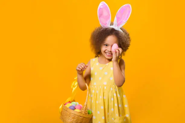Photo of A cheerful Black girl with rabbit ears on her head with a basket of colored eggs in her hands.