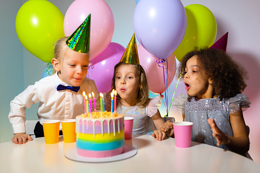 Kids birthday party. Children blowing candles on cake together, vivid Birthday at home. Party decorated with baloons in Festive pastel rainbow theme