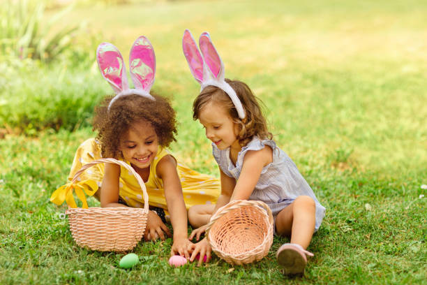 two girls are sitting on the green lawn during easter egg hunt and putting easter eggs in baskets - rabbit hunting imagens e fotografias de stock