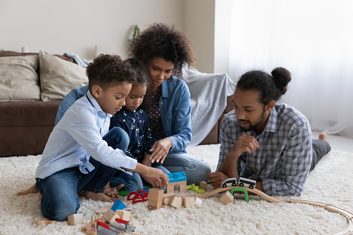 Happy Afro American young parents and two kids constructing building wooden blocks, road, toy railroad model on heating carpeted floor, playing games for imaginations, creative skills development