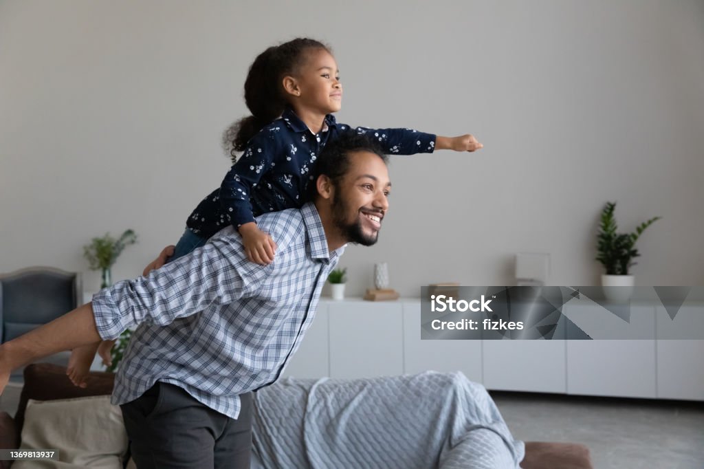 Happy African American dad piggybacking excited proud daughter kid Happy African American dad piggybacking excited proud daughter kid, playing flying superhero, reaching arm forward. Cheerful girl riding fathers back playing active game with daddy at home Father Stock Photo