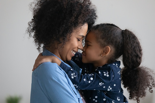 Happy loving Afro American mother holding cute daughter in arms, embracing kid with closed eyes. Smiling mom and sweet little girl hugging with love, affection, gratitude, face touching. Motherhood