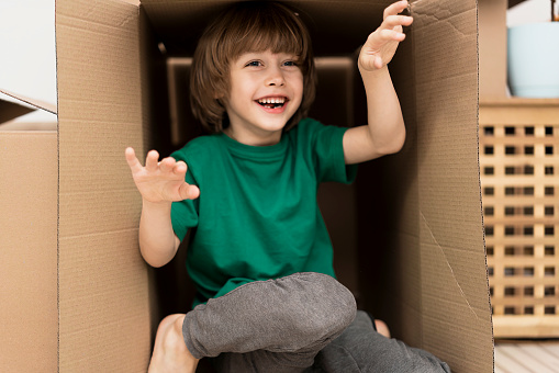 Boy hiding in inside a huge cardboard box. He is playing and looking out of a box. Kid is happy about moving into a new home.