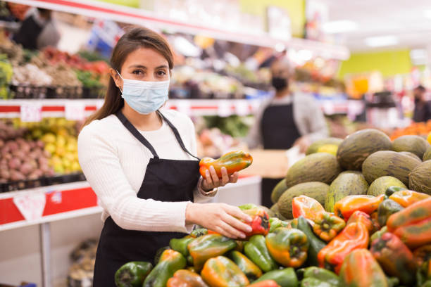 Supermarket worker  in fruit and vegetables section during COVID-19 Portrait of hispanic woman wearing uniform and medical mask standing   in supermarket merchandiser stock pictures, royalty-free photos & images