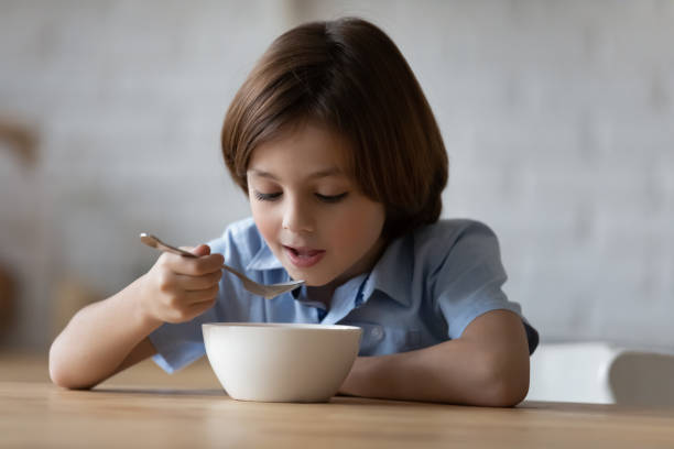 Little boy sit in kitchen holds spoon eating porridge