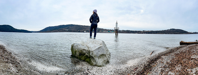 Eurasian young woman at pristine Cates Park, on a cloudy, cold winter day.  A marine terminal for oil is on the far shore in the background.  Dried leaves and seaweed on beach.  North Vancouver, British Columbia, Canada.