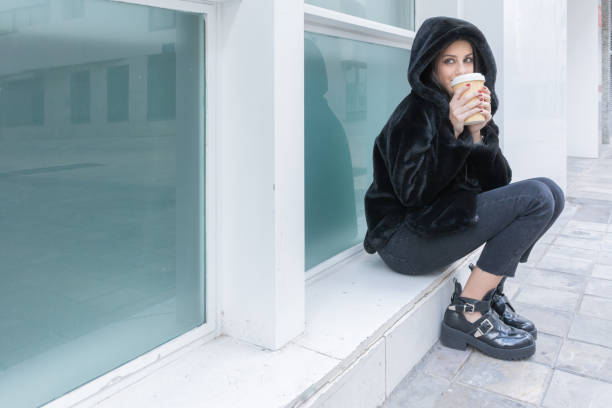 Girl with black coat drinking a take away coffee in a reusable cup sitting on the street