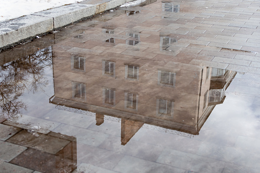 Blurred reflection of residential building in melt water puddle on  sidewalk of city street