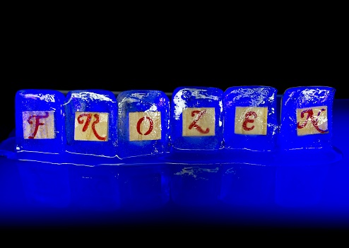 The word frozen spelled out in ice cubes illuminated using a light painting technique. A concept image on a black background with reflection useful for any idea on cold temperatures or frozen. Could be used for weather stories, relationship problems or even financial concepts such as frozen assets.