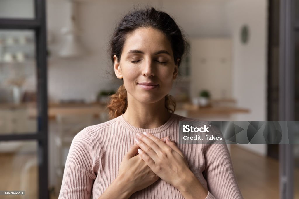 Calm young woman hold hands on chest praying Close up of calm young Caucasian woman hold hands on heart chest feel grateful and thankful. Happy millennial female show gratitude, love and care, pray or visualize. Religion, faith concept. Mental Health Stock Photo