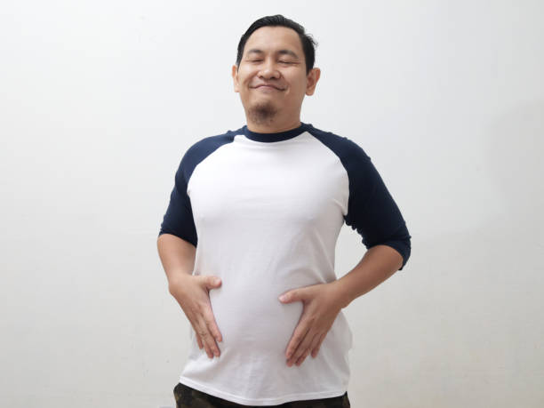 Funny Asian man closing eyes and smiling while holding his distended belly, fully satisfied expression after eating delicious food stock photo