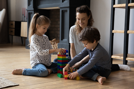 Happy millennial mother or nanny playing toys with cheerful little kids boy girl sitting on warm heated wooden floor in modern living room, constructing building with plastic blocks, playtime activity