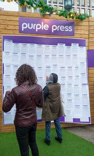 Edinburgh, Scotland, UK - 16th August 2019: Two visitors to the main Underbelly site at Edinburgh Festival Fringe studying reviews of shows on the Purple Press notice board.