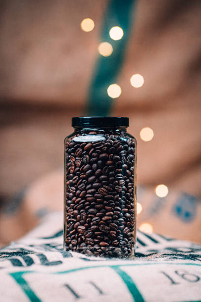 studioaufnahme eines glasmaurglases mit mittelgerösteten kaffeebohnen auf einem jute-kaffeebeutel mit hübschen lichterketten im hintergrund - string coffee bean sack bag stock-fotos und bilder