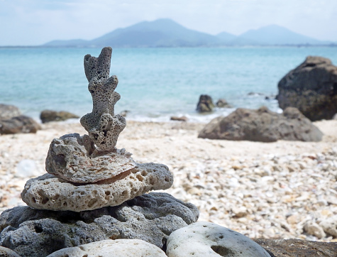 Coral and stones are balanced in a tower on a rocky tropical beach with the ocean and distant mountains visible in the background.