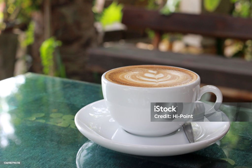 Single cup of coffee on a garden table Hot latte with froth art in a white cup and saucer on a wooden glass-topped coffee table outside in a garden setting on a summer's day. No people. Cappuccino Stock Photo