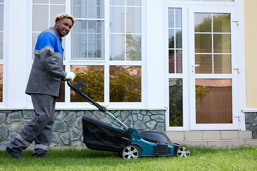 Smiling African man in overalls mows the green grass in a modern garden with a lawn mower. A black man in coveralls pushes a lawnmower in the backyard. Professional lawn care service.
