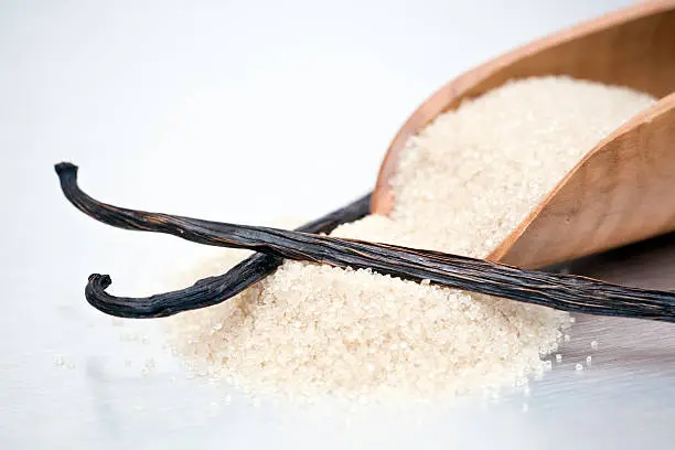 Closeup of processed brown cane sugar in a wooden scoop and two vanilla beans or pods.