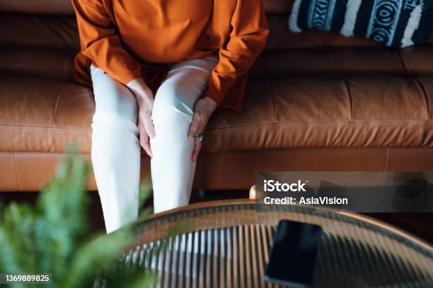 Cropped Shot Of A Distraught Senior Asian Woman Feeling Unwell Suffering From Pain In Leg While Sitting On Sofa In The Living Room At Home Elderly And Health Issues Concept Stock Photo - Download Image Now