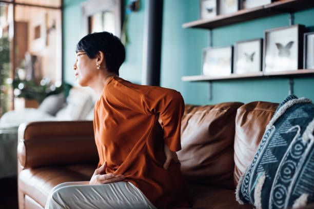 istock A distraught senior Asian woman feeling unwell, suffering from backache, massaging aching muscles while sitting on sofa in the living room at home. Elderly and health issues concept 1369889829