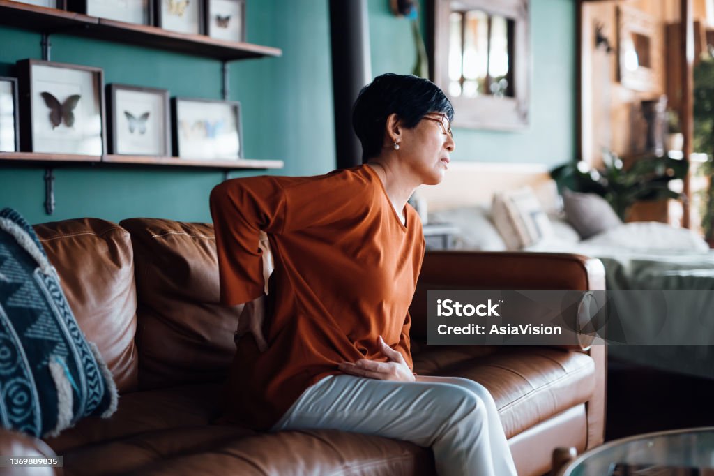 A distraught senior Asian woman feeling unwell, suffering from backache, massaging aching muscles while sitting on sofa in the living room at home. Elderly and health issues concept Backache Stock Photo