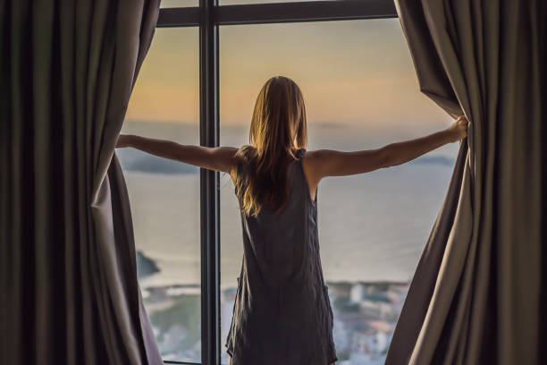 une jeune femme ouvre les rideaux sur une fenêtre avec vue sur la mer - fully unbuttoned photos et images de collection