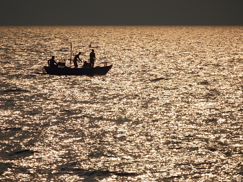 Silhouette of fishermen in a small traditional wooden fishing boat in glistening golden sunset ocean, southeaset Asia.