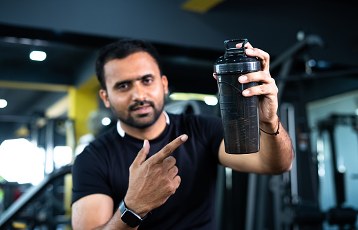 focus on bottle, Confident Indian bodybuilder shacking protein bottle and showing by looking at camera - concept of healthy lifestyle, advertisement and promotion of nutrition supplement.