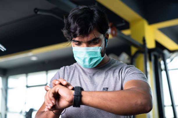 Low angle view of young man with medical face mask checking brunt calories or monitoring hart rate from smartwatch at gym - concept of hygiene, coronavirus covid-19 safety measures and Healthcare. vista de ángulo bajo de un joven con mascarilla médica que revisa las calorías más bajas o monitorea la tasa de hart desde el reloj inteligente en el gimnasio: concpet de higiene, medidas de seguridad covid-19 por coronavirus y atención médica.