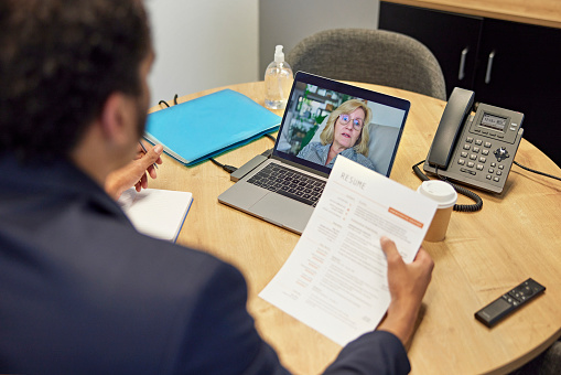 Over the shoulder view of multiracial businessman sitting at table in office and holding résumé as he talks with Caucasian female applicant during video call.
