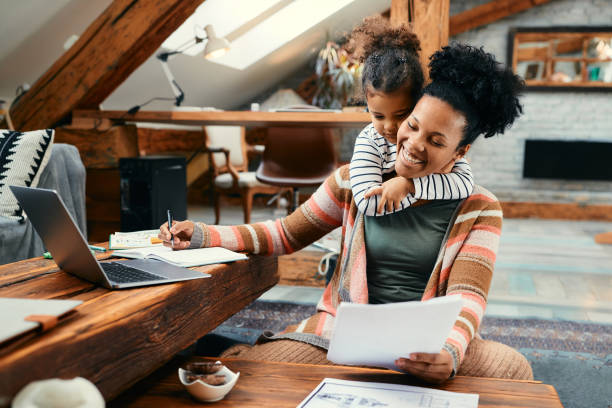 Small black girl embracing her busy mother who is working at home. Happy African American mother using laptop and working on paperwork while her daughter is embracing her at home. home office stock pictures, royalty-free photos & images
