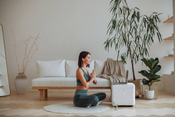 A young woman practices yoga at home in a tracksuit. Sitting in the lotus position A young brunette woman in a tracksuit is doing yoga in an online lesson on a laptop at home in the living room. Sits in the lotus position with closed eyes. In the room there is a white sofa, a white rug, a white pouffe and a house plant tree in a pot yoga lotus position meditating women stock pictures, royalty-free photos & images