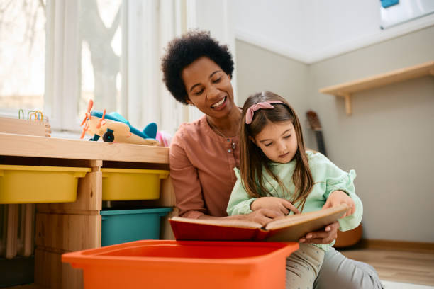 glücklicher afroamerikanischer lehrer und kleines mädchen, das im kindergarten ein buch liest. - child playroom parent indoors stock-fotos und bilder