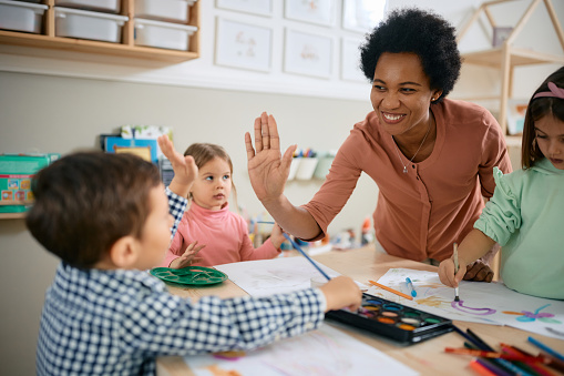 Happy African American teacher and small boy giving high-five during art class at kindergarten.