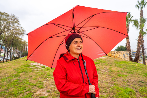 the model, who is 55 years old, has a red raincoat over a beret on her head, green eyes.It's raining indoors.The season is winter.A large umbrella that plays pink in the model's hand.The model is waist-high from the front.portrait.There are palm trees in the park.The model is looking away.He has a high ability to experience positive emotions