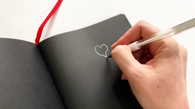 Close-up demonstration of a notebook with black paper for inscriptions in white ink. A woman's hand draws a heart with white ink