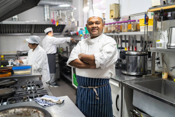 Chef looking at the camera An chef wearing his chef's hat is standing with his arms crossed looking at the camera in a busy industrious kitchen chefs whites stock pictures, royalty-free photos & images