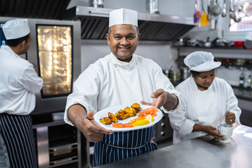 An Indian chef holding a white plate with a freshly cooked chicken appetizer and side salad and smiling at the camera, his colleagues are busy behind him