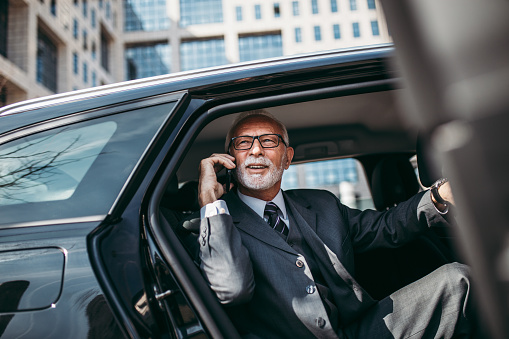Good looking senior business man sitting on backseat in luxury car. He opens car doors and going or stepping out. Big busness bulding in background. Transportation in corporate business concept.