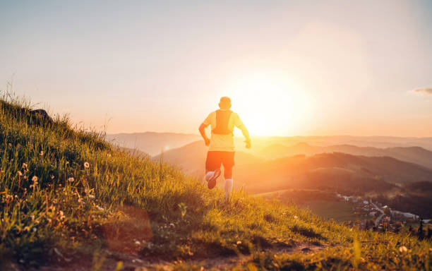 istock Active mountain trail runner dressed bright t-shirt with backpack running endurance marathon race by picturesque hills at sunset time back view photo. Sporty active people backlight concept image. 1369927911