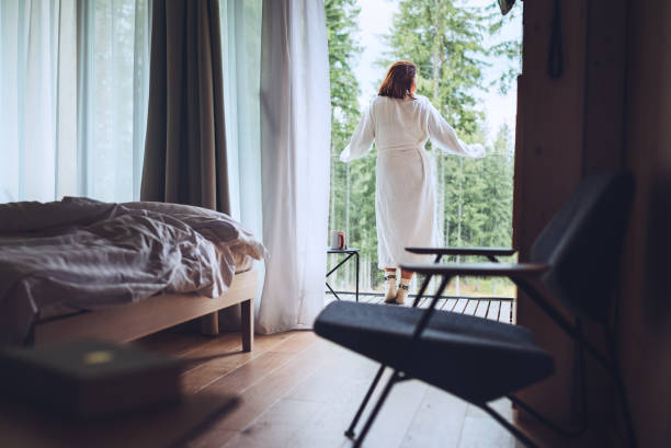 Woman dressed white bathrobe standing on forest house balcony and enjoying fresh air with nature view. Inside the Scandinavian interior design room with a not made bed. Living in wild concept image. stock photo
