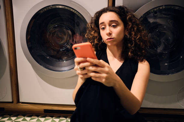 istock Attractive brunette girl thoughtfully using smartphone waiting washing in self-service laundry 1369932439