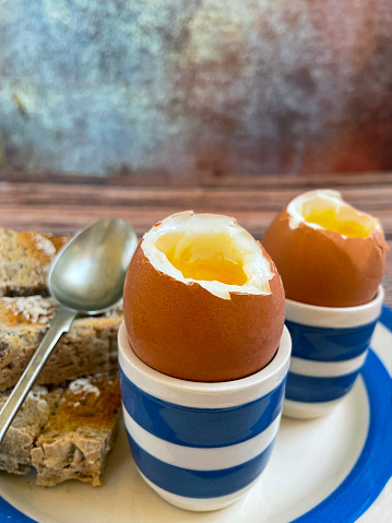 Stock photo showing close-up view of Cornish ware crockery of tea plate and egg cups with wholemeal toast soldiers and topped soft-boiled eggs showing runny yellow yolks.