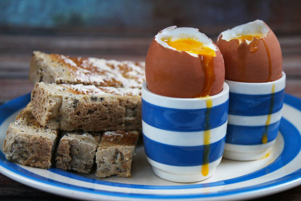 Stock photo showing close-up view of Cornish ware crockery of tea plate and egg cups with wholemeal toast soldiers and topped soft-boiled eggs showing runny yellow yolks.